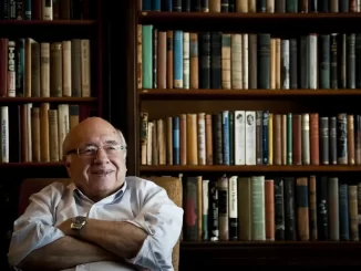 Imagem de Luís Fernando Verissimo sorrindo, sentado em frente a uma estante cheia de livros, durante despedida na Assembleia RS.