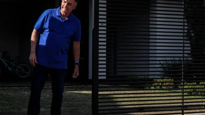 homem-na-frente-de-residencia-noticia-bolsonaro Imagem de um homem em frente a uma residência, com detalhes de uma grade de ferro na porta e ambiente externo, relacionado à notícia sobre o PGR que descartou PF na casa de Bolsonaro e mantém prisão domiciliar do ex-presidente.