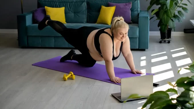 mulher-praticando-exercicios-inicio-bem-estar-online Mulher praticando exercícios físicos em casa, apoiada em mãos e joelhos, assistindo uma aula de bem-estar online na sala com sofá colorido ao fundo.