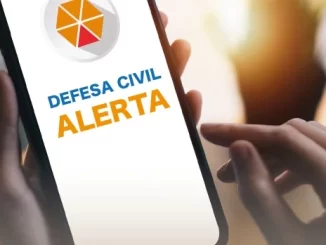 Imagem de uma pessoa segurando um smartphone exibindo o alerta de emergência da Defesa Civil para teste em 13 cidades no sábado.