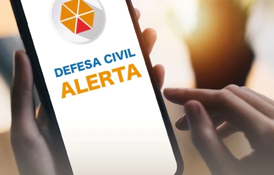 alerta-defesa-civil-teste-emergencia-13-cidades-2023 Imagem de uma pessoa segurando um smartphone exibindo o alerta de emergência da Defesa Civil para teste em 13 cidades no sábado.