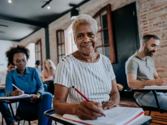 Grupo de adultos participando de aula de alfabetização, evidenciando a importância da alfabetização na inserção no mercado de trabalho formal e no aumento de renda, com destaque para uma senhora idosa sorridente ao centro.