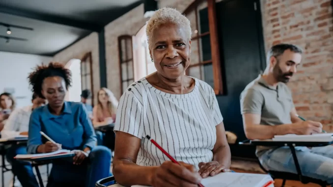 alfabetizacao-de-adultos-aumento-de-renda-trabalho-formal-estudo Grupo de adultos participando de aula de alfabetização, evidenciando a importância da alfabetização na inserção no mercado de trabalho formal e no aumento de renda, com destaque para uma senhora idosa sorridente ao centro.
