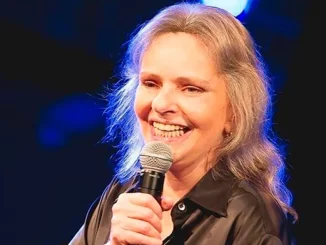 Celebridade Angela Ro Ro sorrindo e segurando um microfone em evento no Rio de Janeiro, aos 75 anos, após luta contra infecção pulmonar.