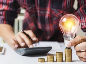 Pessoa segurando uma lâmpada sobre pilhas de moedas, simbolizando economia na energia elétrica, ligado à votação na Câmara que garantirá energia gratuita a 18 milhões de pessoas.