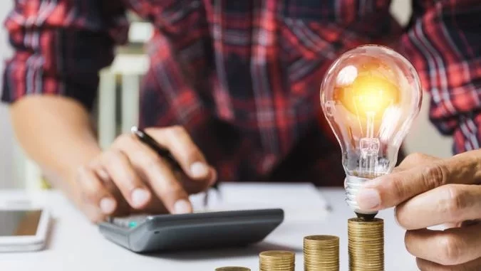 cameravotarampenergiagratuita-jpg Pessoa segurando uma lâmpada sobre pilhas de moedas, simbolizando economia na energia elétrica, ligado à votação na Câmara que garantirá energia gratuita a 18 milhões de pessoas.