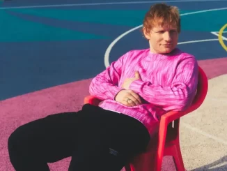Ed Sheeran em ensaio fotográfico com fundo colorido, usando blusa rosa, sentado em cadeira vermelha, preparando-se para temporada nos EUA e promovendo seu lançamento 'Play'