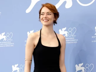 Emma Stone durante o Festival de Veneza 2023, dizendo que acredita em extraterrestres, em destaque na origem da imagem. Ela usa vestido preto e sorri para a câmera, com fundo azul do evento.