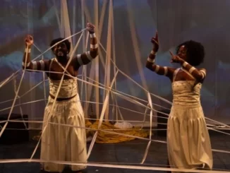 Duas mulheres com pele escura, vestidas com saias longas e camisetas listradas, participando de uma apresentação no Festival Artes Vertentes, interagindo com fios e objetos artísticos.