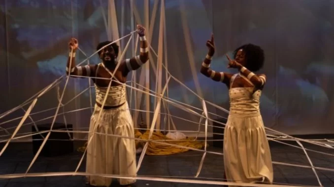 festival-artes-vertentes-musica-danca-arte-jpg Duas mulheres com pele escura, vestidas com saias longas e camisetas listradas, participando de uma apresentação no Festival Artes Vertentes, interagindo com fios e objetos artísticos.