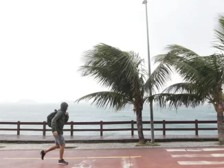 Pessoa caminhando na orla do Rio de Janeiro com ventos fortes de mais de 76 km/h devido à frente fria nesta terça-feira, causando impacto na paisagem e no movimento da praia.