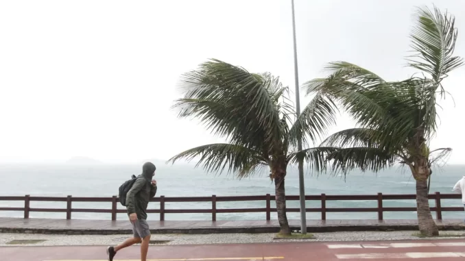 frente-fria-ventos-acima-de-76-kmh-rio-nesta-terca-jpg Pessoa caminhando na orla do Rio de Janeiro com ventos fortes de mais de 76 km/h devido à frente fria nesta terça-feira, causando impacto na paisagem e no movimento da praia.