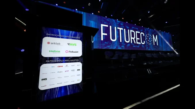 futurecom2025-debate-ia-data-centros-sustentabilidade-brasil Imagem do palco do Futurecom 2025 com telões exibindo debates sobre inteligência artificial, data centers e sustentabilidade no Brasil. Evento conta com patrocinadores como Arkklok, AVS, Intelbras e polkadot, promovendo inovação tecnológica e sustentabilidade no setor de telecomunicações brasileiro.