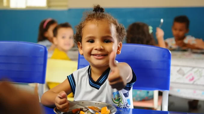 investimento-mundial-em-merenda-alcanca-80-milhoes-de-alunos Criança sorridente em uma escola durante a refeição escolar, representando o aumento do investimento mundial em merenda que chega a 80 milhões de alunos.