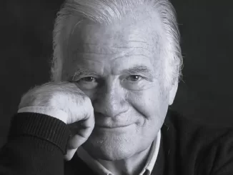 Imagem de Mino Carta, criador da Veja e Carta Capital, sorrindo para a câmera em retrato preto e branco, aos 91 anos, memória de um jornalista influente.