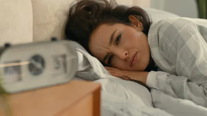 mulher-noites-mal-dormidas-pressao-alta-risco-avc Mulher com expressão de preocupação na cama, com um despertador ao lado, ilustrando os riscos de noites mal dormidas, pressão alta e AVC.
