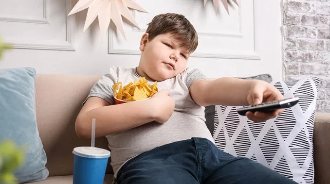obesidade-infantil-unicef-alarme-global Criança observando TV com batatas fritas e refrigerante, ilustrando a preocupação global com o aumento da obesidade infantil, conforme alerta do Unicef.