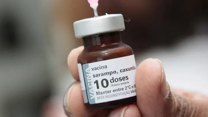 vacina-contra-sarampo-2025-duplicacao-criterio Pessoa segurando uma ampola de vacina contra sarampo, com uma seringa sendo preparada para aplicação, destacando a importância da imunização e o aumento na procura pela vacina em 2025.