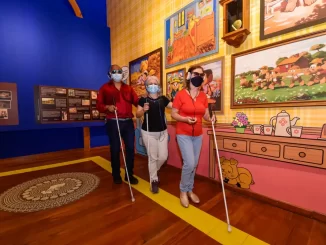 Três pessoas com deficiência visual visitando exposição de artes no Itaú Cultural, com pinturas coloridas ao fundo, usando bengalas e máscaras de proteção.