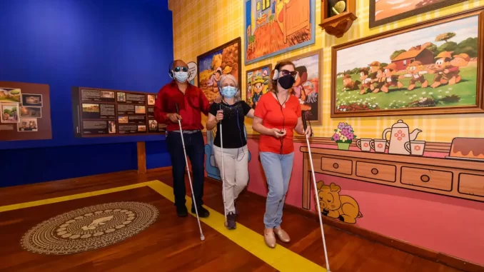 itaucultural-lanca-dossie-acessibilidade-arte Três pessoas com deficiência visual visitando exposição de artes no Itaú Cultural, com pinturas coloridas ao fundo, usando bengalas e máscaras de proteção.