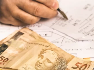 Imagem mostrando uma mão escrevendo em papel com notas, e notas de dinheiro de 50 reais ao lado, relacionada à variação dos juros do consignado entre bancos segundo o Procon-SP.