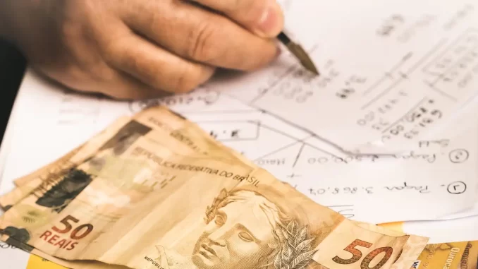 procon-sp-juros-consignado-variam-bancos Imagem mostrando uma mão escrevendo em papel com notas, e notas de dinheiro de 50 reais ao lado, relacionada à variação dos juros do consignado entre bancos segundo o Procon-SP.