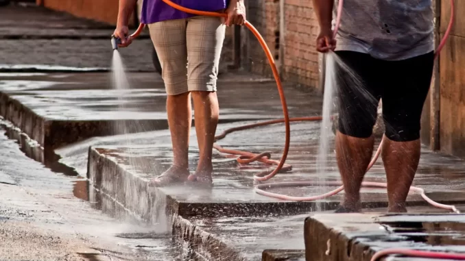 Pessoas lavando calçada com mangueiras em uma rua, destacando o desperdício de água no Brasil, que desperdiça 6 mil piscinas por dia, conforme estudo.