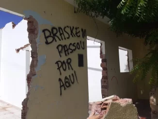 Imagem de uma parede destruída com grafite que diz 'Braskem passou por aqui', relacionada à indenização de R$ 1,2 bilhão por afundamento de solo em Maceió.