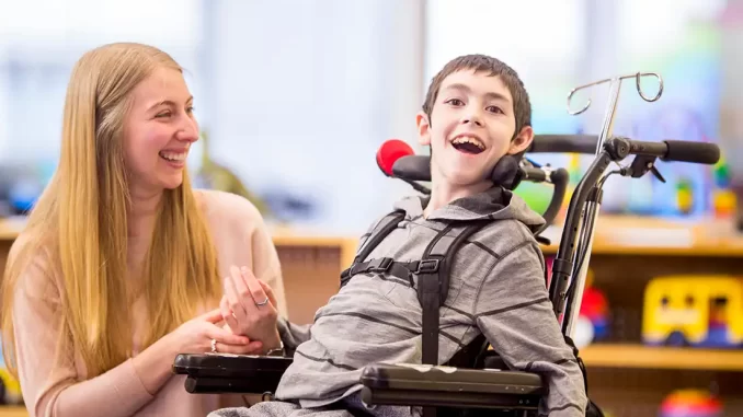 Criança com atrofia muscular espinhal em cadeira de rodas sorrindo ao lado de uma mulher, possivelmente sua mãe, em ambiente de clínica ou hospital após autorização da Anvisa para testar terapia gênica pela Fiocruz contra a Atrofia Muscular Espinhal grave.