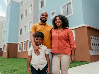 Família feliz na frente de um prédio de habitação popular, celebrando o aumento do teto do Minha Casa Minha Vida pelo Conselho do FGTS.