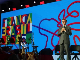 Macron vestido de terno no palco do Festival Cidades do Futuro em Salvador, com painel colorido ao fundo destacando elementos de arte e ativismo.