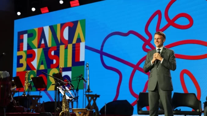 festival-cidades-do-futuro-salvador-arte-ativismo Macron vestido de terno no palco do Festival Cidades do Futuro em Salvador, com painel colorido ao fundo destacando elementos de arte e ativismo.