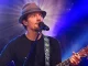 Jason Mraz em performance ao vivo confirmando turnê sul-americana e cinco shows no Brasil, tocando guitarra e cantando em palco iluminado.
