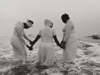 Foto de três mulheres negras de roupas brancas deitados na praia, segurando as mãos na água, simbolizando o legado da escravidão e reafirmando a importância da memória histórica na reabertura do Museu Histórico Nacional com mostra internacional.