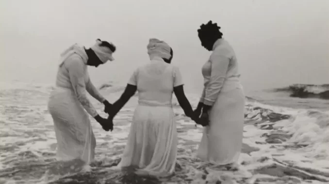 Foto de três mulheres negras de roupas brancas deitados na praia, segurando as mãos na água, simbolizando o legado da escravidão e reafirmando a importância da memória histórica na reabertura do Museu Histórico Nacional com mostra internacional.