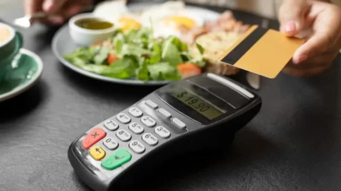 Pessoa usando cartão de débito para pagar um almoço em um restaurante, com uma maquininha de pagamento e alimentos à sua frente, simbolizando benefícios do novo decreto