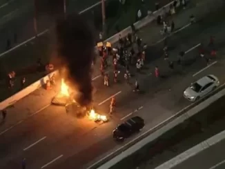 Protesto na rua com barricadas queimadas, fogo e pessoas ao redor, relacionados à discussão sobre o Senado tipificar bloqueio de vias como novo crime.
