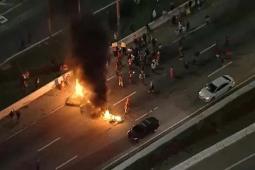 Protesto na rua com barricadas queimadas, fogo e pessoas ao redor, relacionados à discussão sobre o Senado tipificar bloqueio de vias como novo crime.