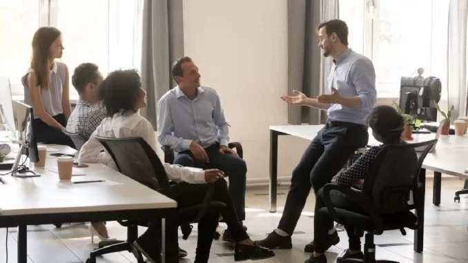 Reunião de equipe em uma empresa nos EUA, com gerentes sendo cortados e equipes sendo ampliadas por chefe, refletindo tendências empresariais atuais.