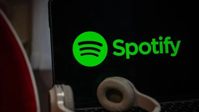 spotify-processado-streams-falsos-drake Imagem mostra logo do Spotify na tela de um laptop, com fone de ouvido na frente, indicando a polêmica sobre o processamento de streams falsos de Drake.