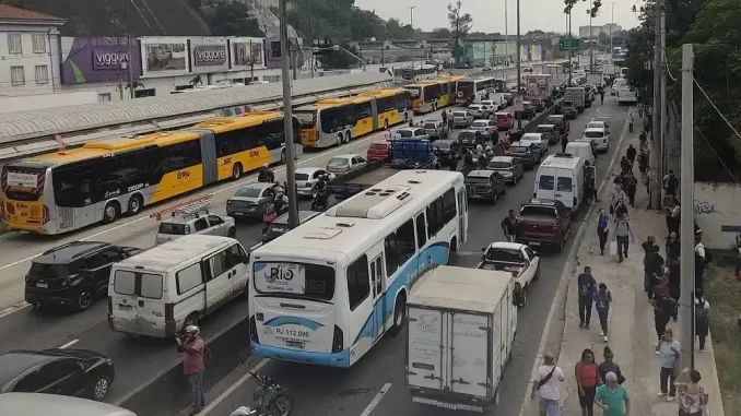 Tiroteio na Avenida Brasil no Rio de Janeiro causa bloqueio temporário, com trânsito congestionado, ônibus parados e pessoas caminhando na calçada, após troca de tiros.