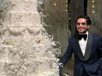 Confeiteiro dos Famosos Denilson Lima em evento com bolo decorado elaborado, usando terno preto com lapela de cetim, durante divulgação de áudio familiar em vídeo