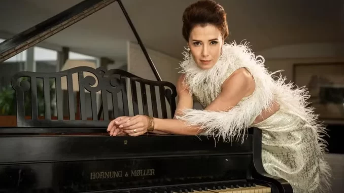 Cena do episódio 5 da série 'Ângela Diniz' mostrando uma atriz em clima de tensão extrema ao lado de um piano, retratando a morte da personagem, com foco na expressão facial de drama e suspense.