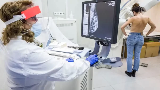 exame-mamografia-qualidade-de-vida-cancer Profissional de saúde realizando exame de mamografia, uma mulher em fundo observando a imagem na tela, em ambiente clínico com equipamentos médicos.