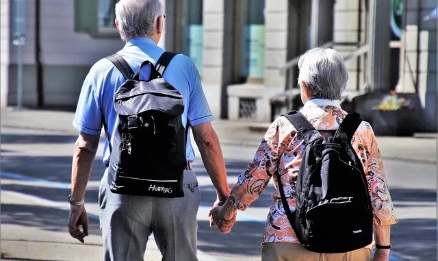 habitos-de-vida-para-viver-ate-100-anos-bem-jpg Casal de idosos caminhando na rua com mochilas, ilustrando hábitos de vida saudável para viver até 100 anos bem, inspirado em estudo sobre hábitos de centenários.