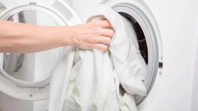 importancia-de-lavar-lencois-travesseiros-edredoes-microbiologa Pessoa colocando roupas de cama, travesseiros e edredons na máquina de lavar, ilustrando a importância de lavar esses itens corretamente, de acordo com uma microbióloga.