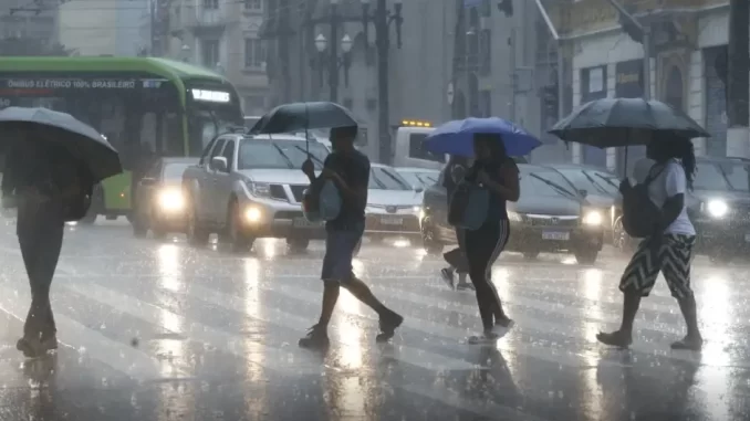 inmet-alerta-vermelho-tempestades-ventos-100kmh Pessoas com guarda-chuvas atravessando a rua durante tempestade forte devido ao alerta vermelho do Inmet para tempestades e ventos de 100 km/h