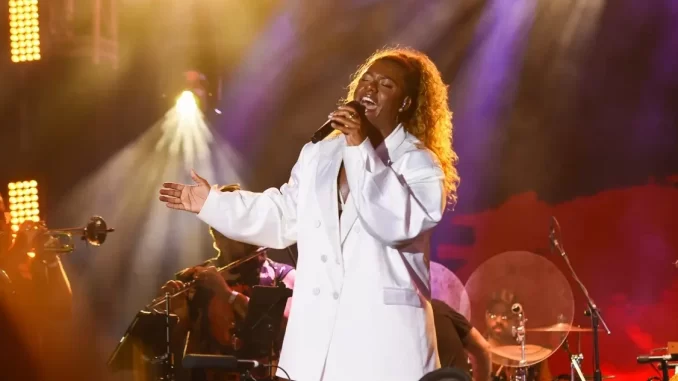 liniker-turne-bye-bye-caju-quatro-capitais-brasileiras-jpg Liniker se apresentando em show da turnê BYE BYE CAJU em uma das quatro capitais brasileiras, com iluminação de palco vibrante e banda ao fundo.