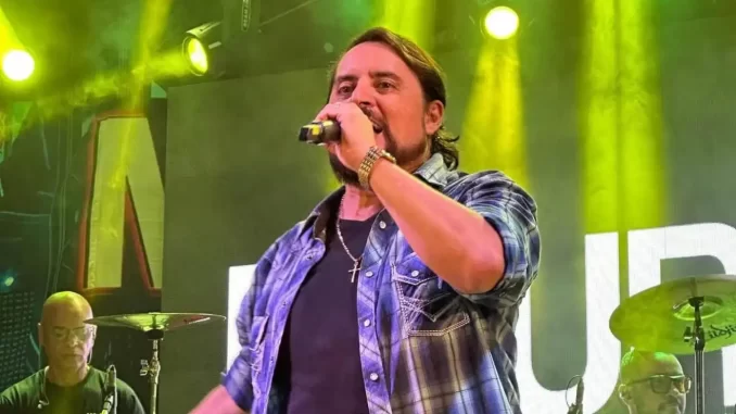 mauri-irmao-de-chitaozinho-e-xororo-morre-em-acidente-rodoviario Mauri, irmão de Chitãozinho e Xororó, morto em acidente rodoviário, durante apresentação musical com iluminação amarela ao fundo.