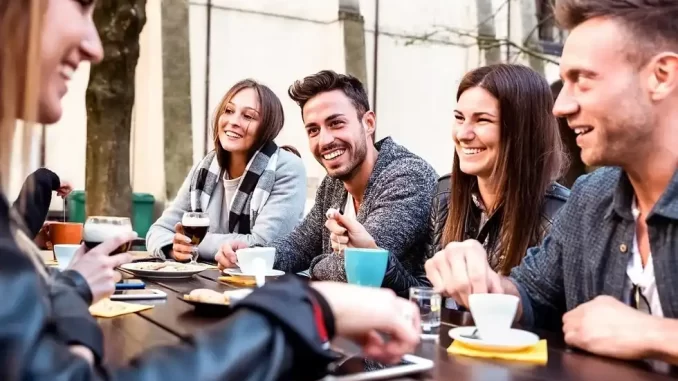 Grupo de jovens millennials sorrindo e conversando ao redor de uma mesa ao ar livre, celebrando sucesso financeiro, com destaque para o relatório que indica que essa geração pode ser a mais rica.